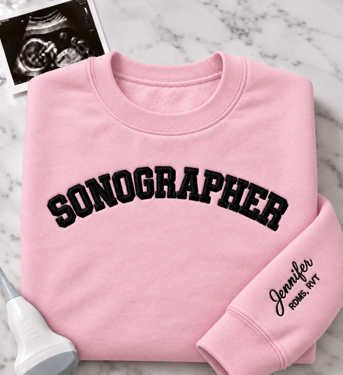 A folded light pink crewneck sweatshirt with 'SONOGRAPHER' embroidered across the chest in black arched lettering, and a customizable name & credentials embroidered on the sleeve. styled on a white marble background with an ultrasound photo and wand props.