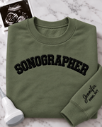 An olive green crewneck sweatshirt with 'SONOGRAPHER' embroidered across the chest in black arched lettering, and a customizable name & credentials embroidered on the sleeve. styled on a white marble background with an ultrasound photo and wand props.