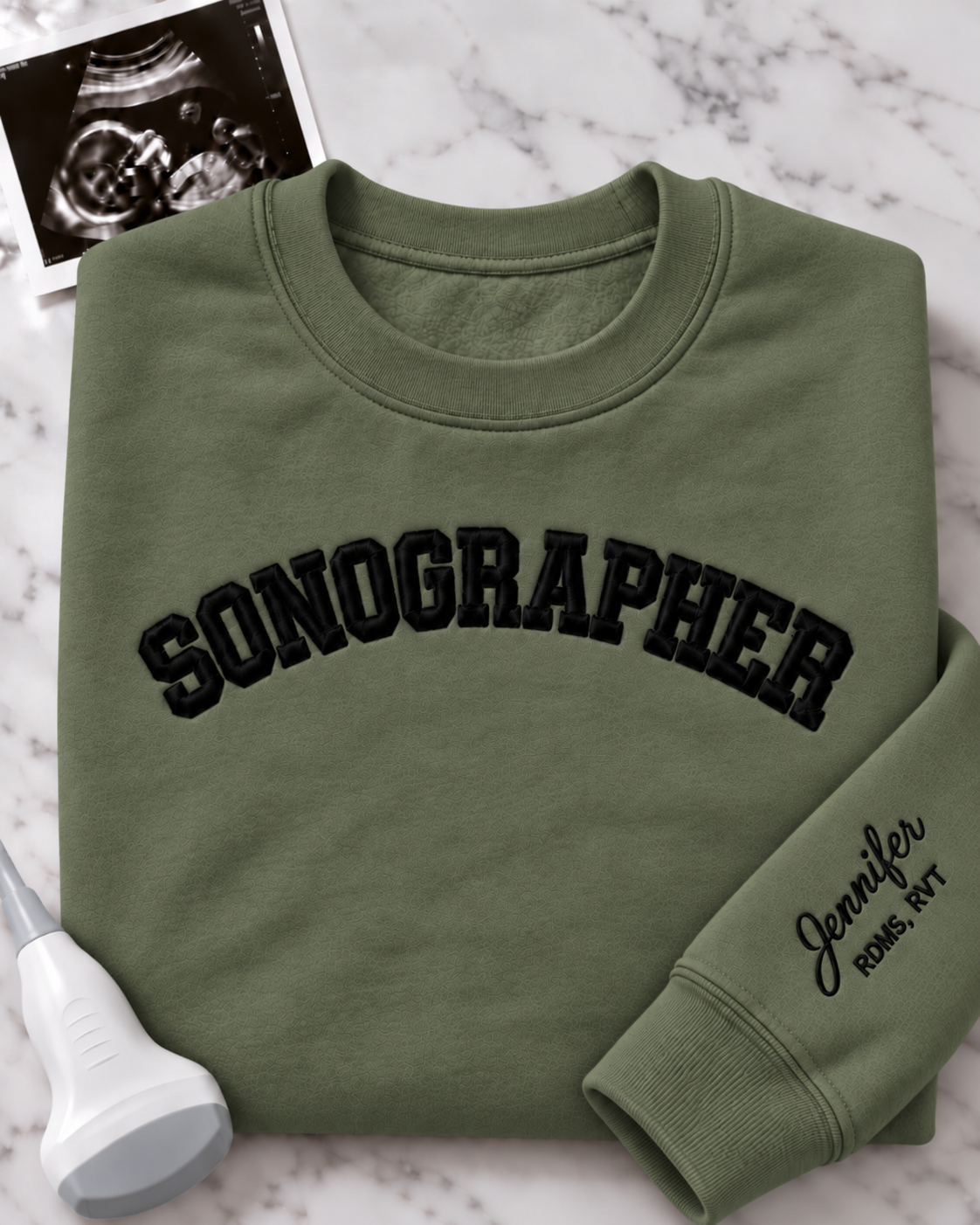 An olive green crewneck sweatshirt with 'SONOGRAPHER' embroidered across the chest in black arched lettering, and a customizable name & credentials embroidered on the sleeve. styled on a white marble background with an ultrasound photo and wand props.