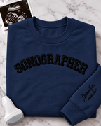 A navy blue crewneck sweatshirt with 'SONOGRAPHER' embroidered across the chest in black arched lettering, and a customizable name & credentials embroidered on the sleeve. styled on a white marble background with an ultrasound photo and wand props.