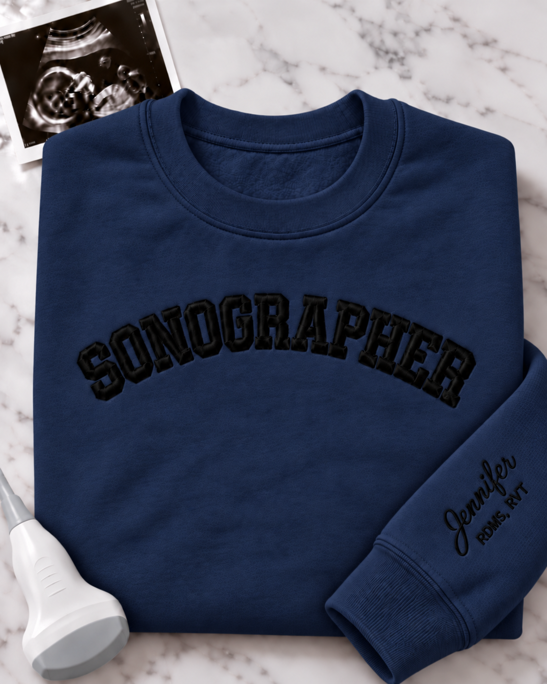 A navy blue crewneck sweatshirt with 'SONOGRAPHER' embroidered across the chest in black arched lettering, and a customizable name & credentials embroidered on the sleeve. styled on a white marble background with an ultrasound photo and wand props.
