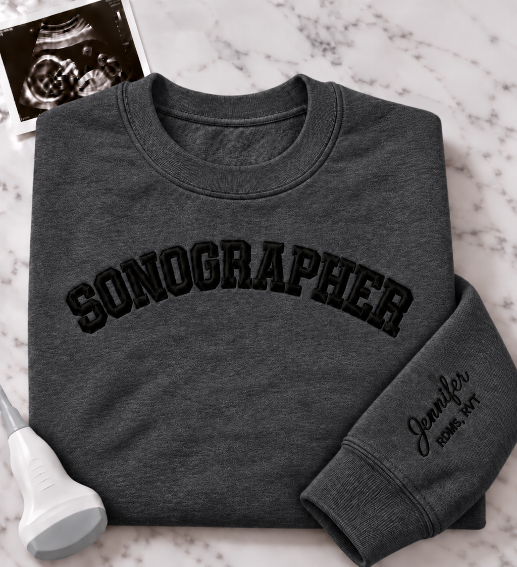 A folded dark gray crewneck sweatshirt with 'SONOGRAPHER' embroidered across the chest in black arched lettering, and a customizable name & credentials embroidered on the sleeve. styled on a white marble background with an ultrasound photo and wand props.