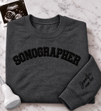A folded dark gray crewneck sweatshirt with 'SONOGRAPHER' embroidered across the chest in black arched lettering, and a customizable name & credentials embroidered on the sleeve. styled on a white marble background with an ultrasound photo and wand props.