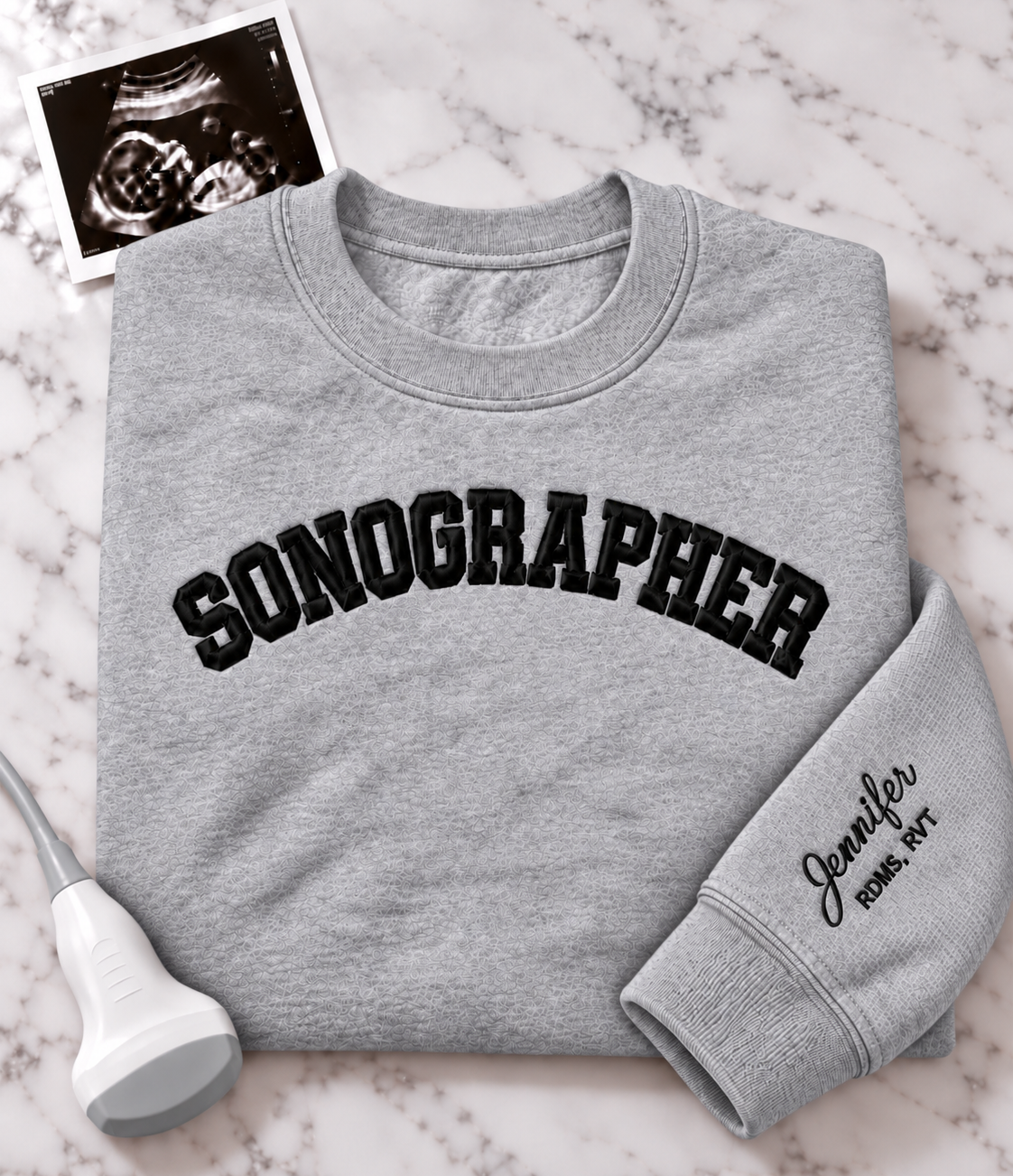 A folded light gray crewneck sweatshirt with 'SONOGRAPHER' embroidered across the chest in black arched lettering, and a customizable name & credentials embroidered on the sleeve. styled on a white marble background with an ultrasound photo and wand props.