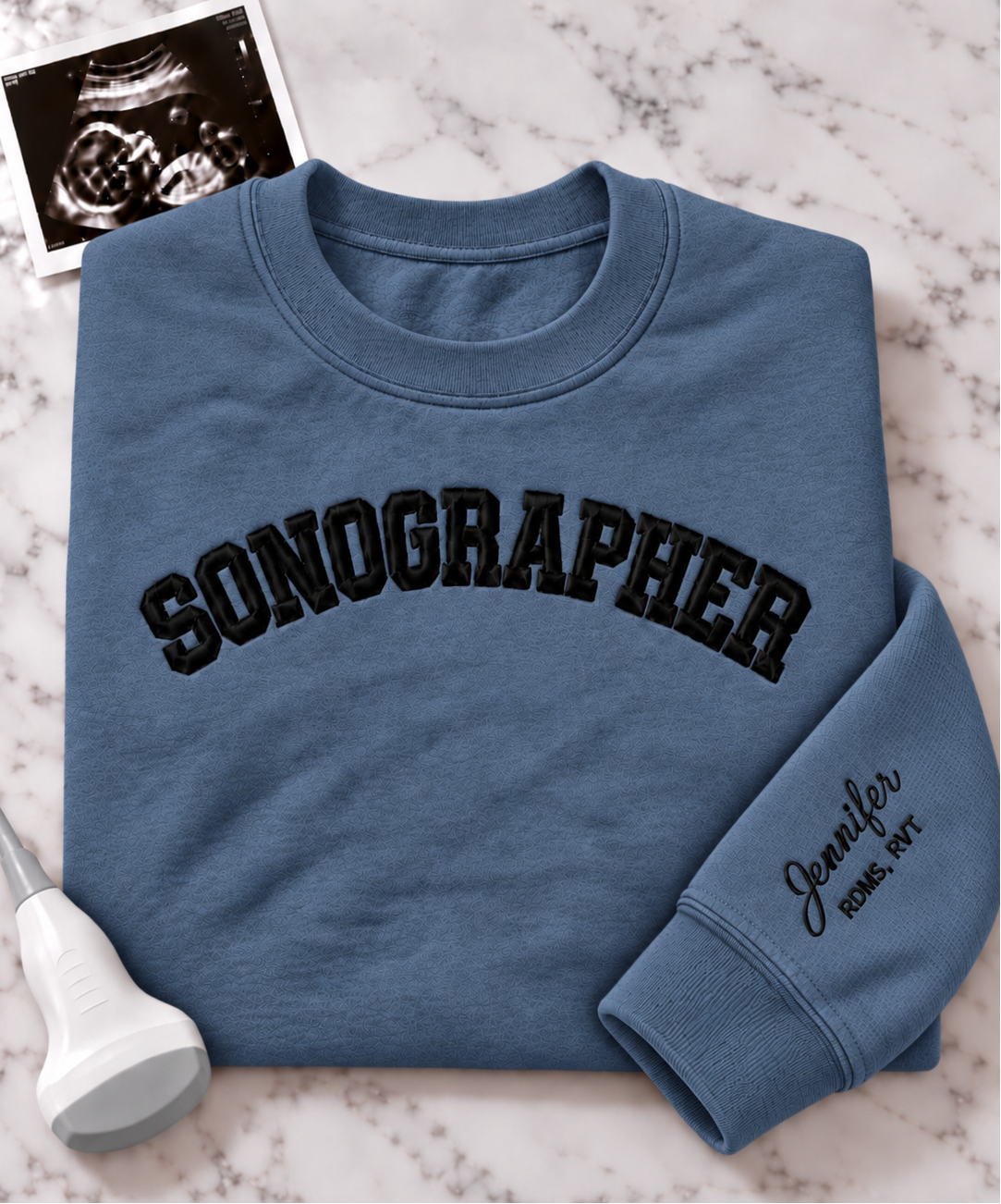 A folded dusty blue crewneck sweatshirt with 'SONOGRAPHER' embroidered across the chest in black arched lettering, and a customizable name & credentials embroidered on the sleeve. styled on a white marble background with an ultrasound photo and wand props.